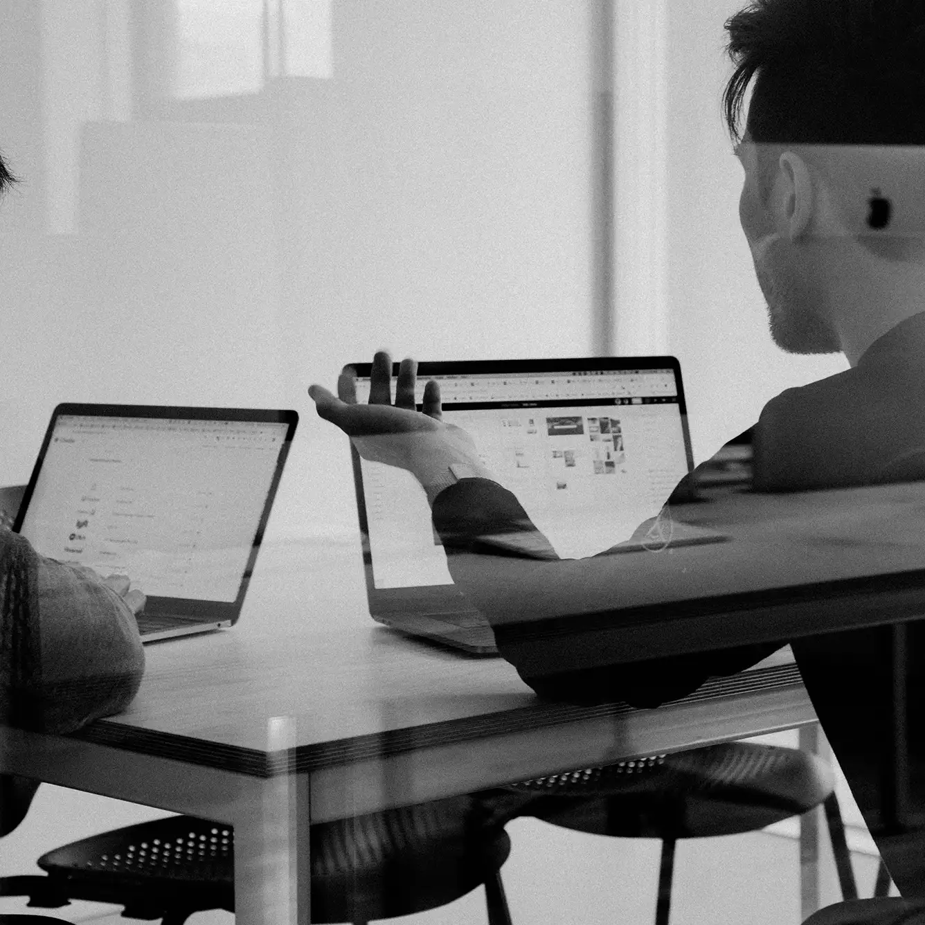 Two people working on laptops at a table, one person gesturing with their hand while discussing something.