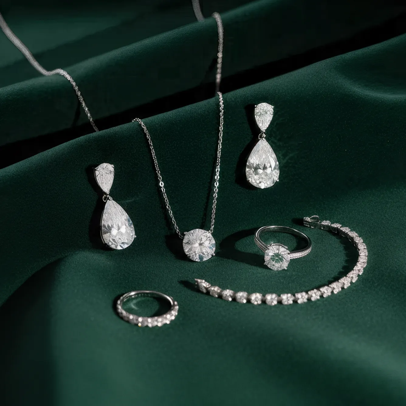 Collection of silver jewelry including teardrop earrings, a round pendant necklace, two rings, and a diamond bracelet on dark green fabric.