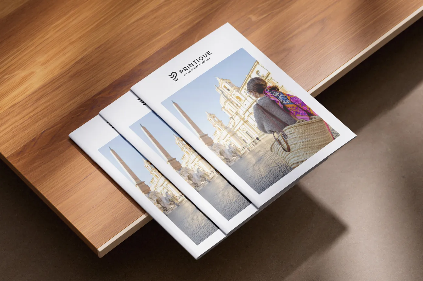 Three Printique catalogs with a cover photo of a woman with a colorful scarf and woven bag standing in a historic square with an obelisk and classical buildings.