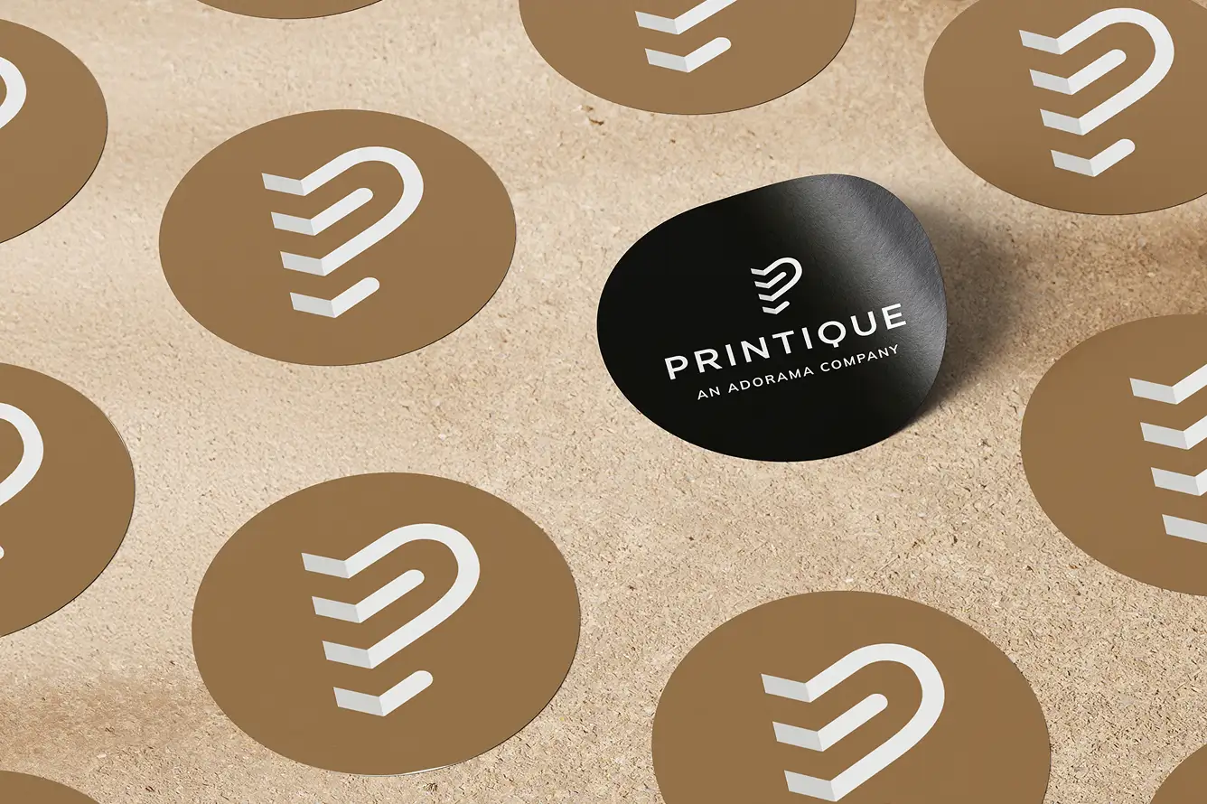 Black circular sticker with white Printique logo and text 'PRINTIQUE AN ADORAMA COMPANY' among multiple brown circular stickers with a white logo on a textured beige surface.