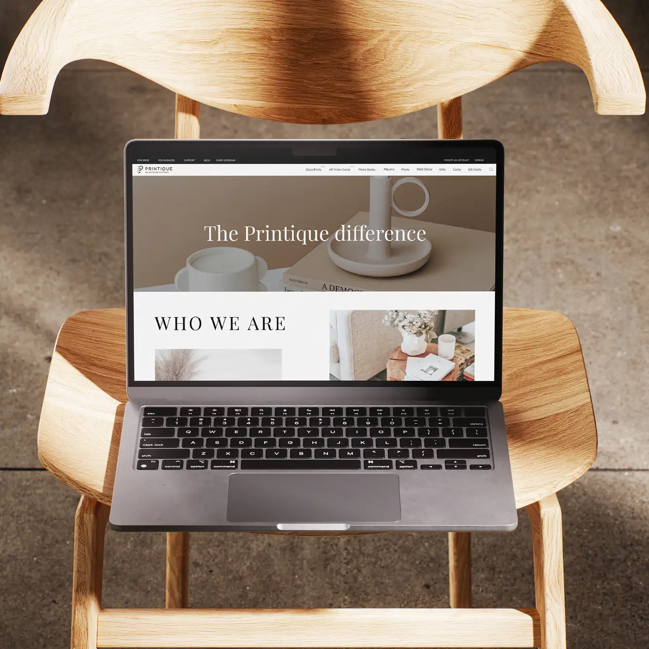 Laptop displaying Printique website on a wooden chair with text 'The Printique difference' and 'Who We Are'.