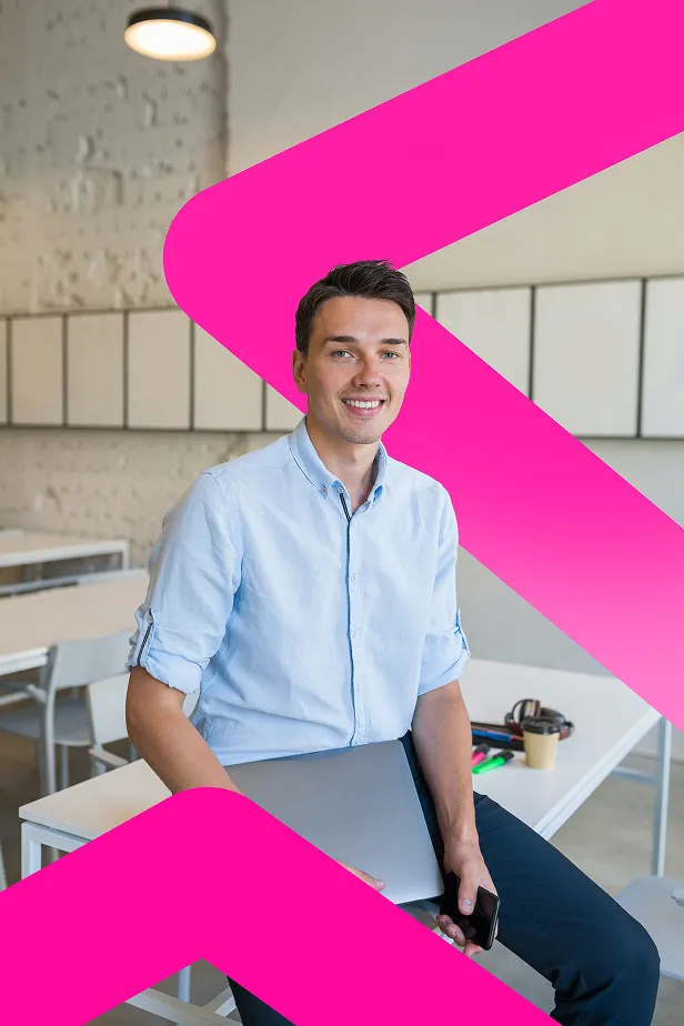 Smiling young man in a light blue shirt holding a laptop and smartphone while sitting in a modern office.