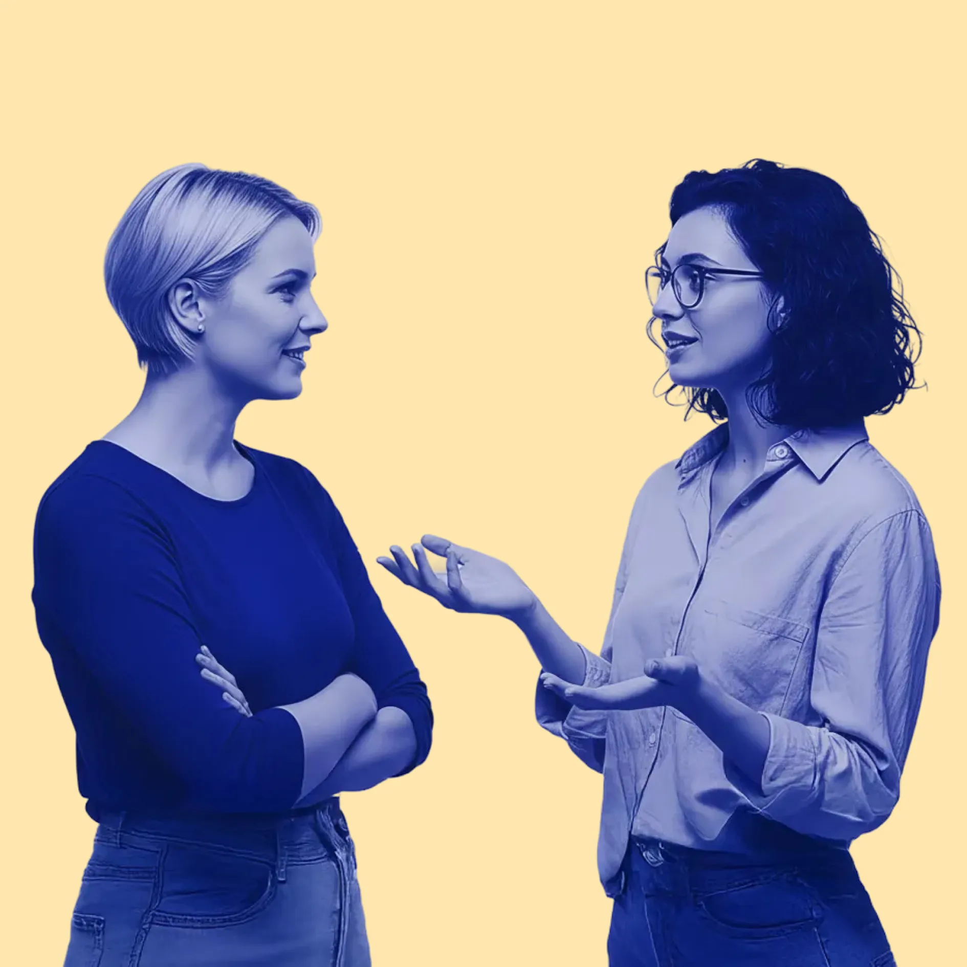 Two women engaged in conversation, one with short blonde hair and arms crossed, the other with glasses and curly dark hair gesturing with her hands.