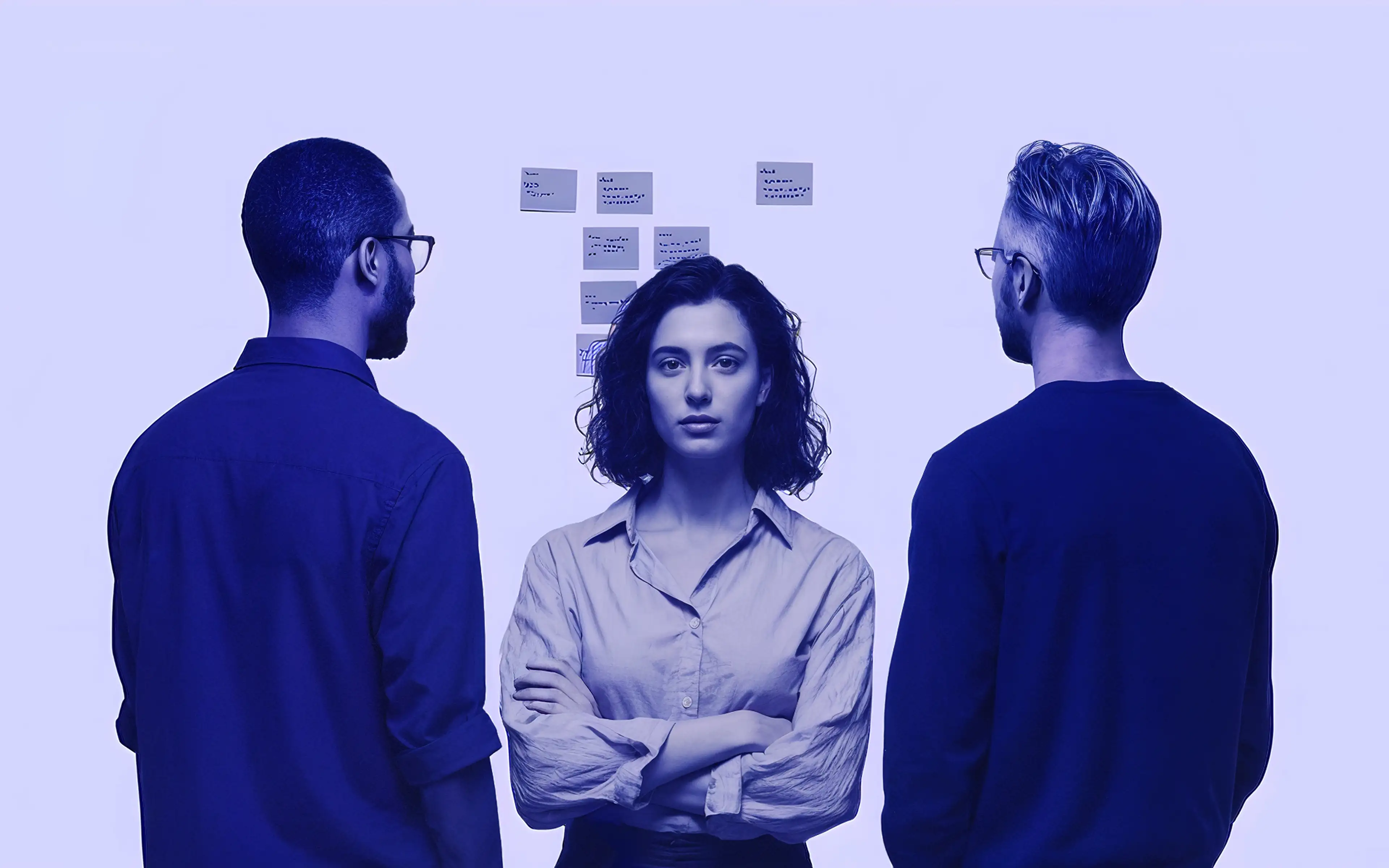 Three professionals standing and looking at sticky notes on a wall, with a woman facing forward and two men facing sideways.
