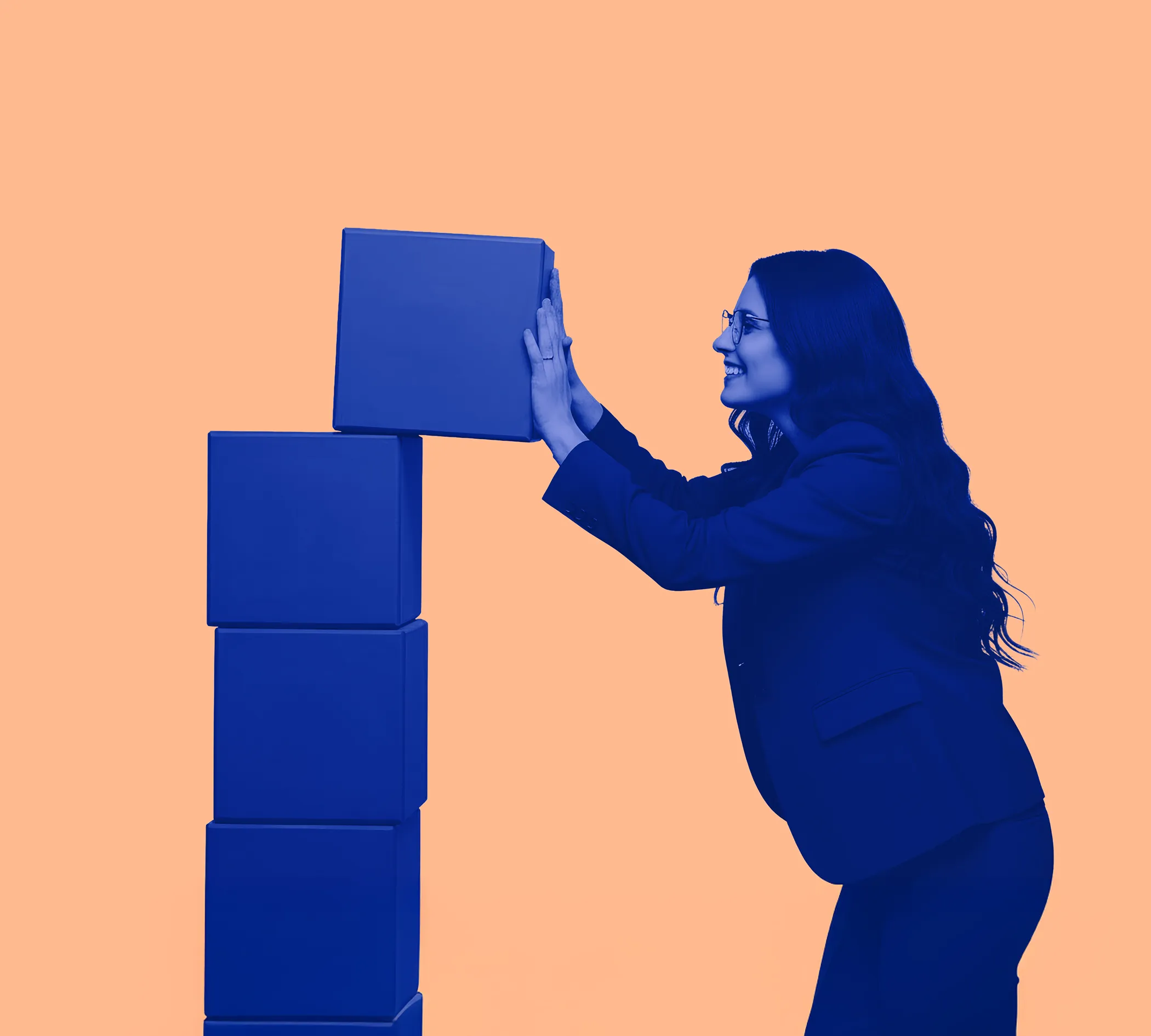 Woman in glasses and business attire placing a cube on top of a stack of cubes against a peach background.