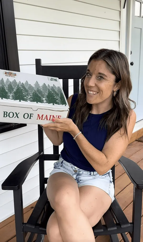 Katie Zarrilli in a navy sleeveless top and denim shorts sitting on a black rocking chair, holding a white box with green pine tree prints labeled 'BOX OF MAINE'.
