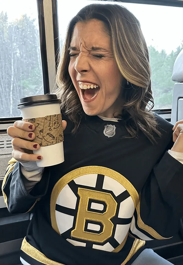 Katie Zarrilli wearing a Boston Bruins hockey jersey holding a coffee cup and expressing excitement or joy.