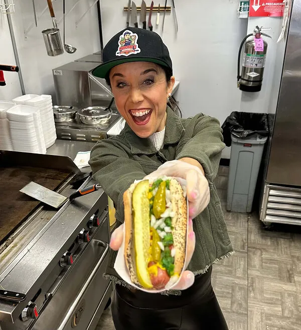 Katie Zarrilli wearing a baseball cap and gloves holding a hot dog topped with pickles, onions, and relish in a commercial kitchen.