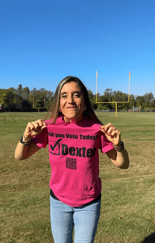 Katie Zarrilli on a football field holding up a pink shirt that says 'Did you Vote Today? Dexter' with a checkmark.