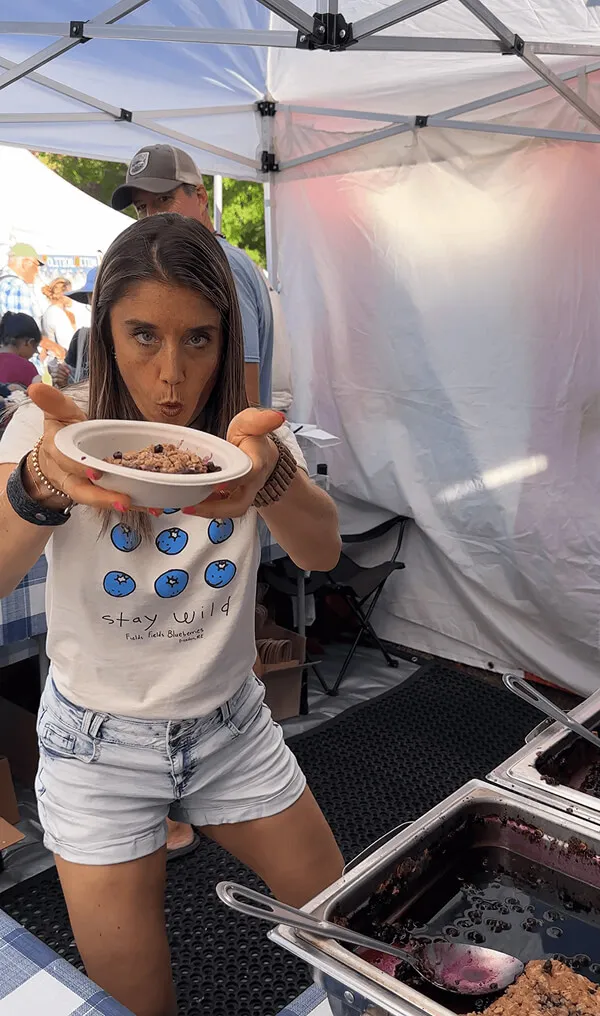 Katie Zarrilli in a 'stay wild' blueberry shirt holding a plate of blueberry oatmeal towards the camera under a white tent.