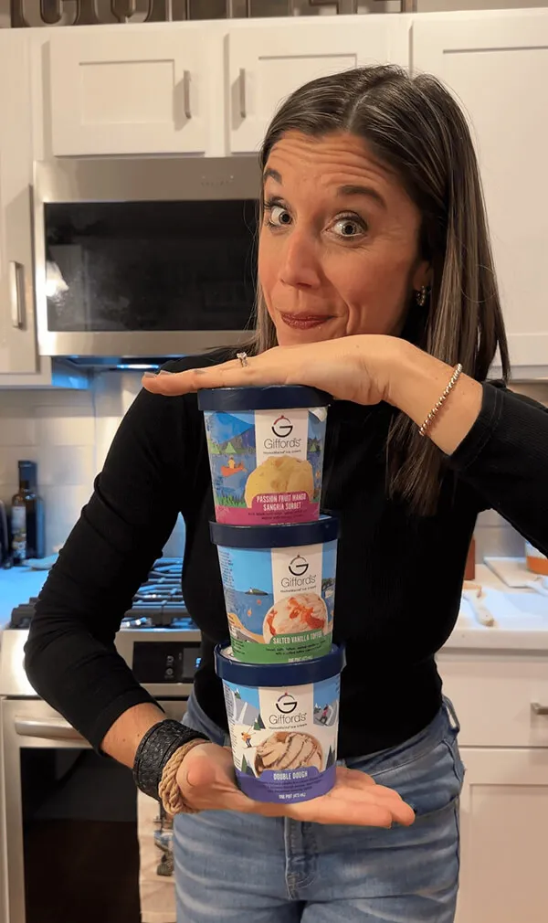 Katie Zarrilli in black shirt holding and playfully presenting a tower of three Gifford's ice cream containers in a kitchen.