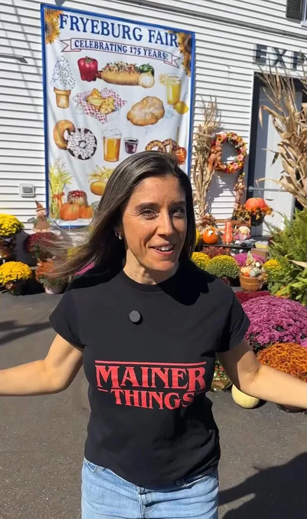 Katie Zarrilli wearing a black 'Mainer Things' t-shirt standing outdoors near fall decorations and a Fryeburg Fair banner.