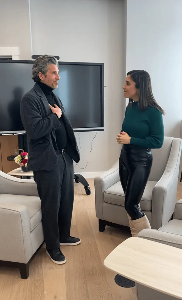 Katie Zarrilli and Patrick Dempsey standing and talking in a modern office lounge.