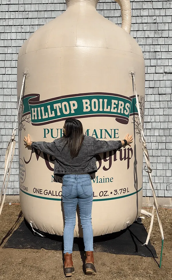 Katie Zarrilli hugging a giant inflatable container of Hilltop Boilers Pure Maine Maple Syrup outdoors.