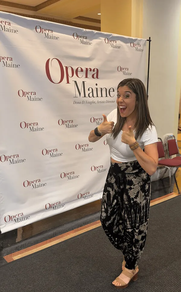 Katie Zarrilli in a white top and patterned black pants smiling and giving two thumbs up beside an Opera Maine banner.