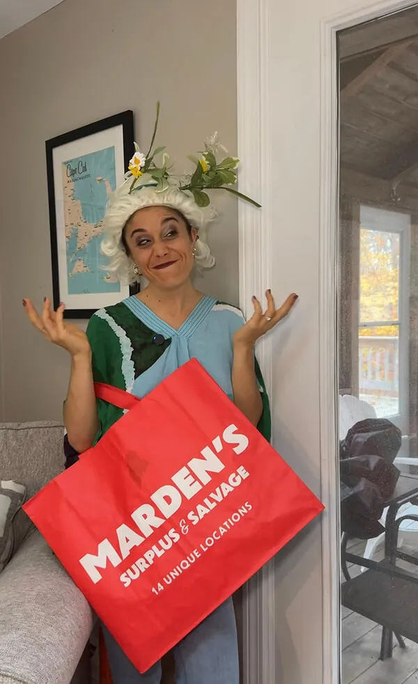 Katie Zarrilli wearing a floral wreath and a wig, shrugging while holding a red Marden's Surplus & Salvage shopping bag.