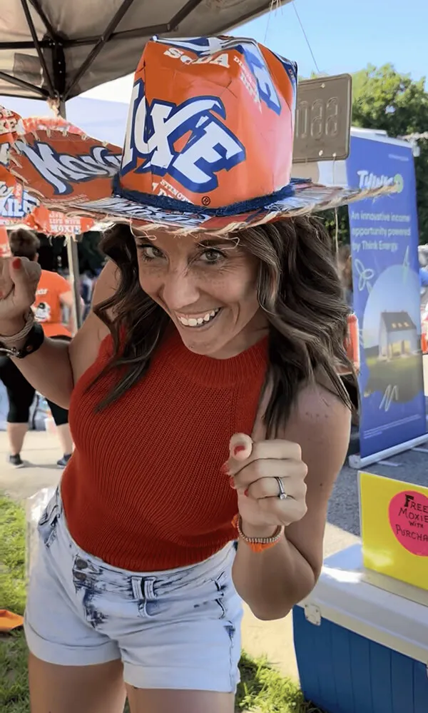 Katie Zarrilli wearing a red sleeveless top and white shorts, posing energetically with a colorful orange and blue novelty hat at an outdoor event.