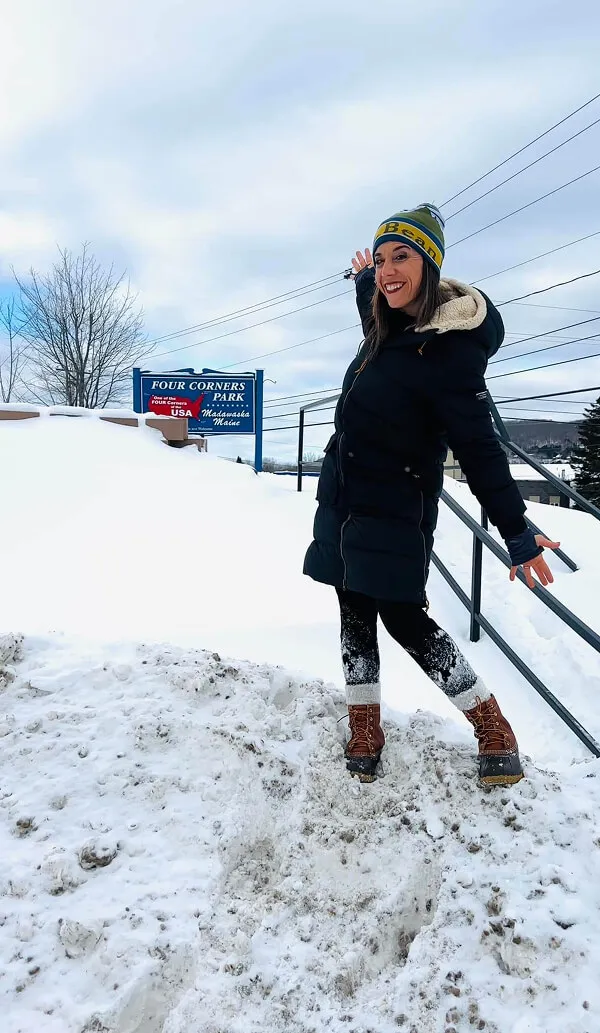Katie Zarrilli in winter coat, boots, and hat posing happily on snow near Four Corners Park sign in Madawaska, Maine.