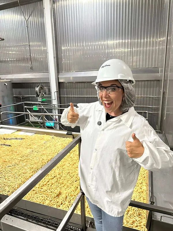 Katie Zarrilli in a white lab coat, safety glasses, hairnet, and hard hat giving two thumbs up beside a conveyor belt full of fries in a food processing facility.