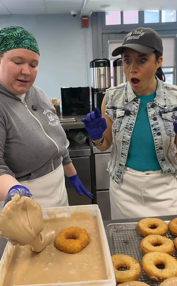 Katie Zarrilli with a women at a kitchen counter; one dips a donut into batter while the other watches with a surprised expression.