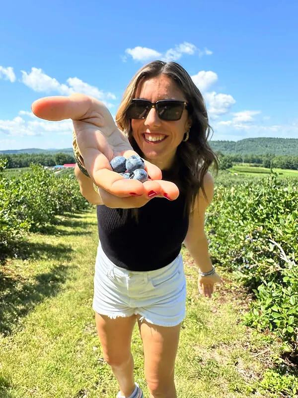 Katie Zarrilli in sunglasses holding out handful of blueberries in a sunny blueberry field.