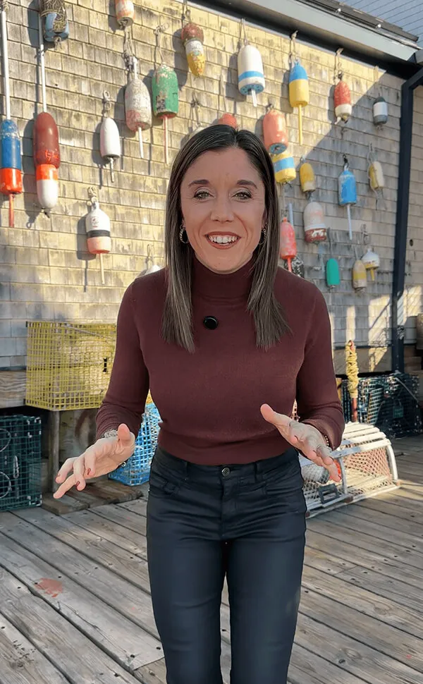 Katie Zarrilli wearing a maroon turtleneck and black pants standing on a wooden deck with colorful buoys hanging on a shingled wall behind her.