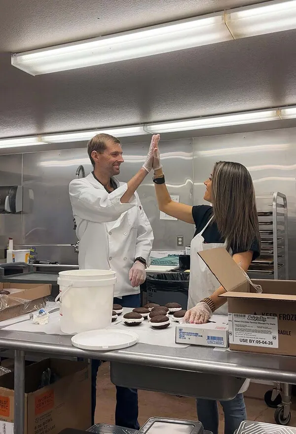 Katie Zarrilli and her husband wearing gloves and aprons give each other a high five in a commercial kitchen with baked chocolate cupcakes on the counter.