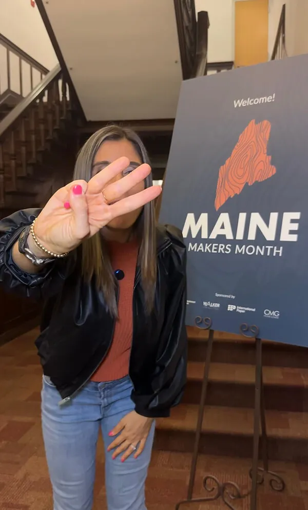 Katie Zarrilli wearing a black jacket and jeans holding up three fingers in front of a Maine Makers Month welcome sign.