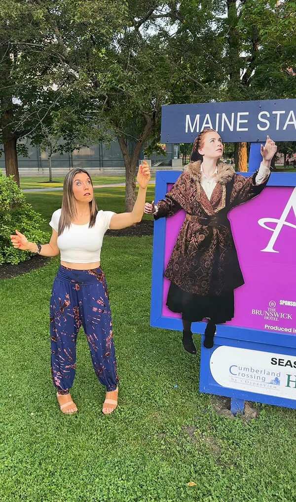 Katie Zarrilli in white top and patterned blue pants mimicking the pose of a historical figure in a coat on a Maine State sign outdoors.