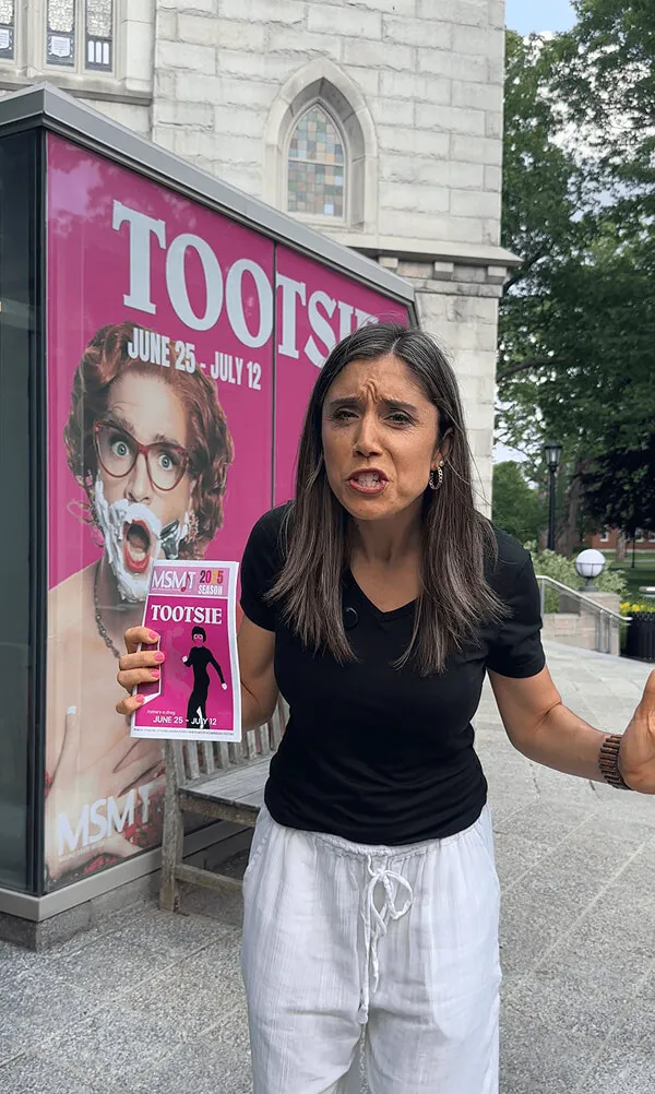 Katie Zarrilli in black shirt and white pants standing outdoors holding a Tootsie playbill with a Tootsie poster displayed behind her.