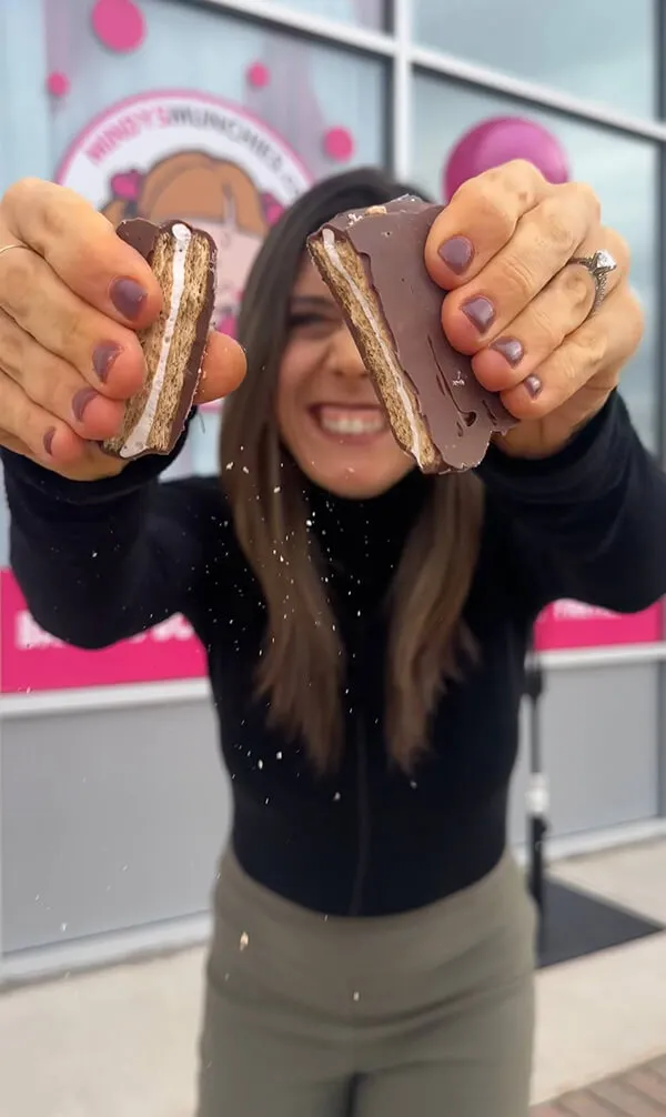 Katie Zarrilli smiling and holding a broken chocolate-covered marshmallow treat close to the camera.