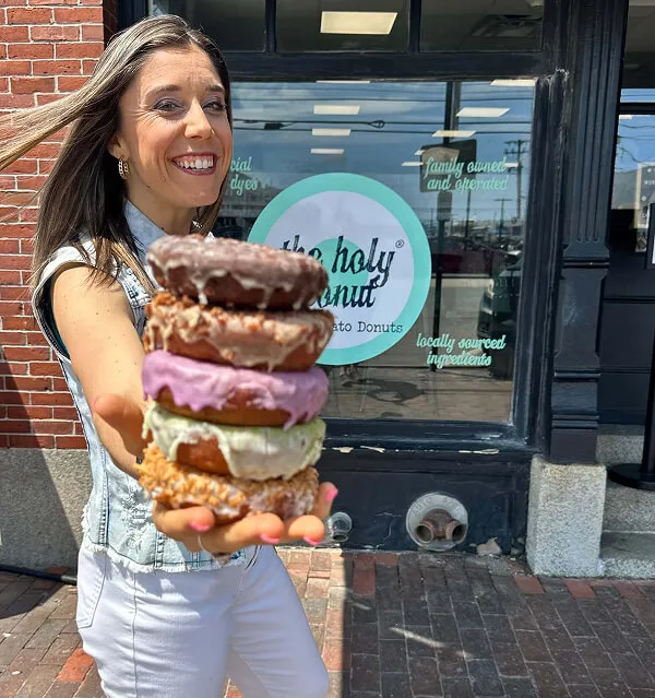 Katie Zarrilli holding a stack of colorful donuts in front of a shop window with The Holy Donut logo.