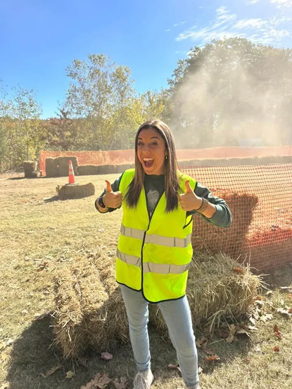 Katie Zarrilli wearing a neon yellow safety vest giving two thumbs up outdoors near hay bales and orange construction fencing under a clear blue sky.
