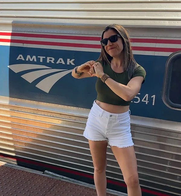 Katie Zarrilli wearing sunglasses and a green crop top makes a heart shape with her hands in front of an Amtrak train.