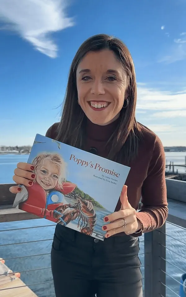 Katie Zarrilli smiling and holding a children's book titled 'Poppy's Promise' with an illustration of a child wearing a red life jacket and holding a lobster.