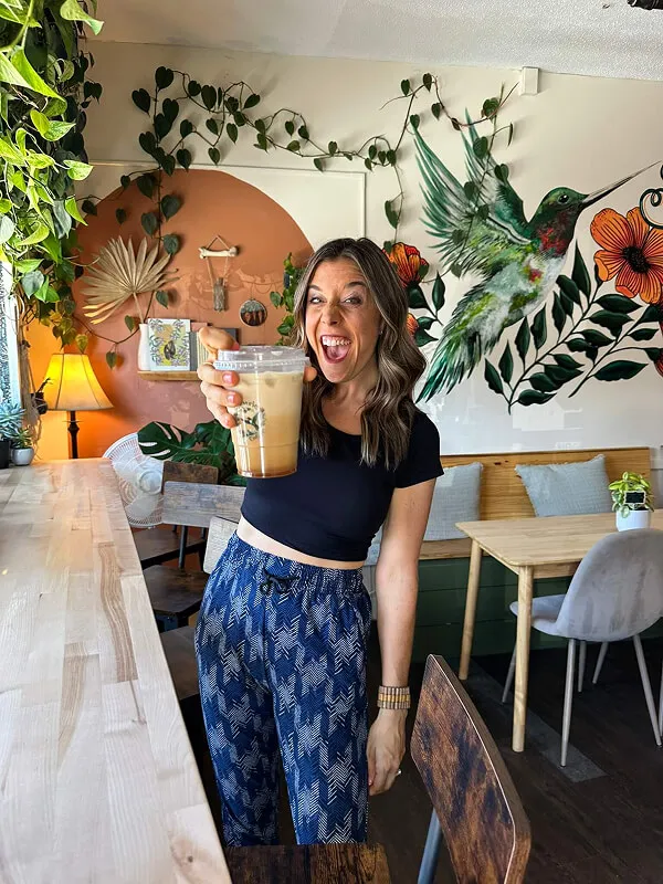 Katie Zarrilli in a black crop top and blue patterned pants holding up a cup of iced coffee in Reverie Coffee with a hummingbird mural and plants.