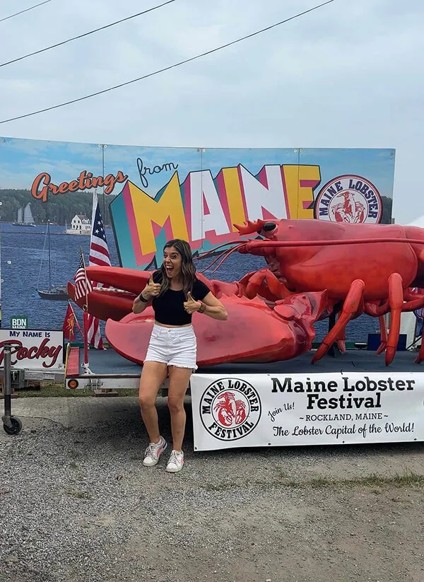 Katie Zarrilli  giving thumbs up stands in front of a giant red lobster and a sign reading 'Maine Lobster Festival, Rockland, Maine, The Lobster Capital of the World!'