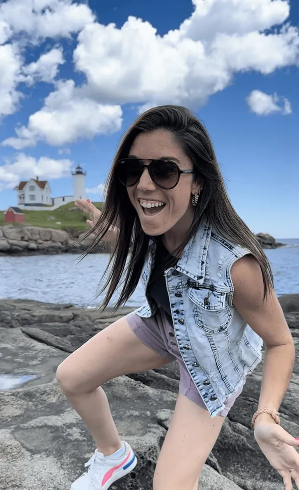 Katie Zarrilli wearing sunglasses, denim vest, and sneakers smiling and posing on rocks near water with a lighthouse and house in the background under a blue sky with clouds.