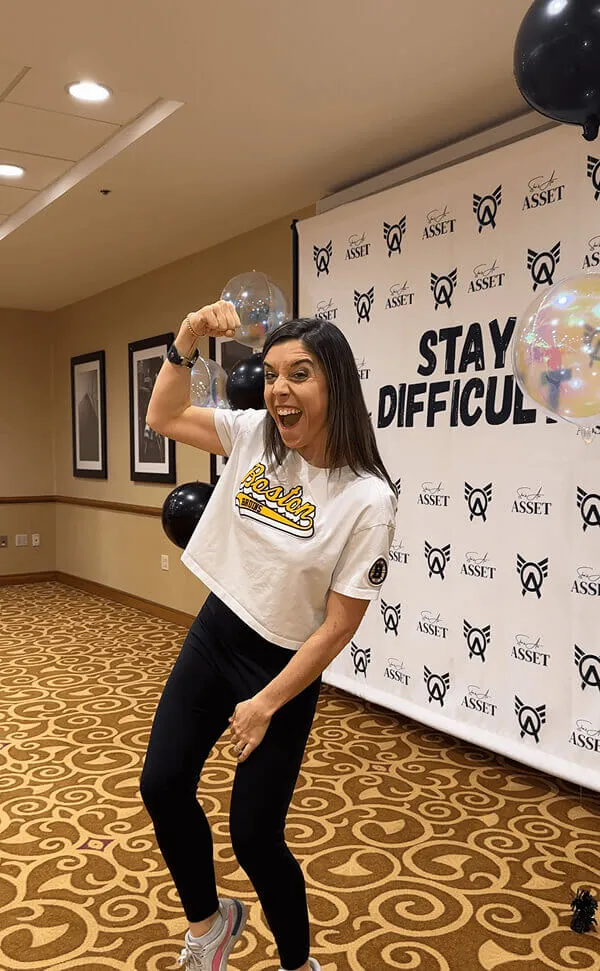 Katie Zarrilli in a white Boston Bruins crop top flexing her arm and smiling in a decorated indoor space with balloons and a banner saying 'Stay Difficult'.