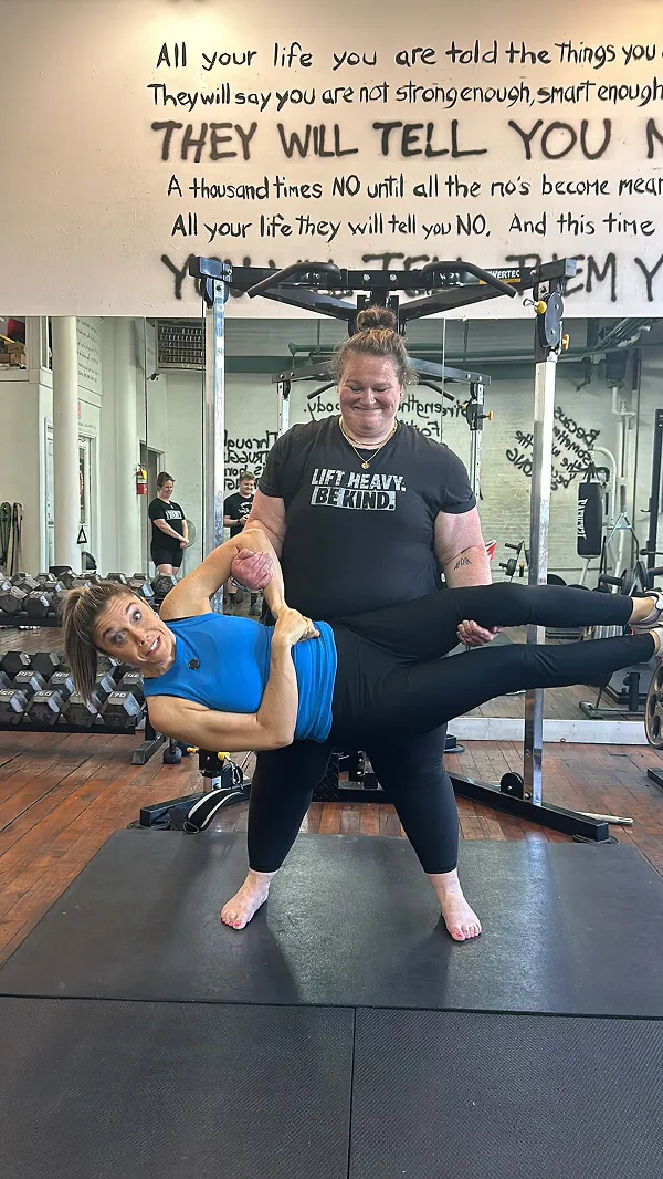 Katie Zarrilli at a gym doing a partnered exercise; one woman in black T-shirt lifting the other in blue tank top horizontally.