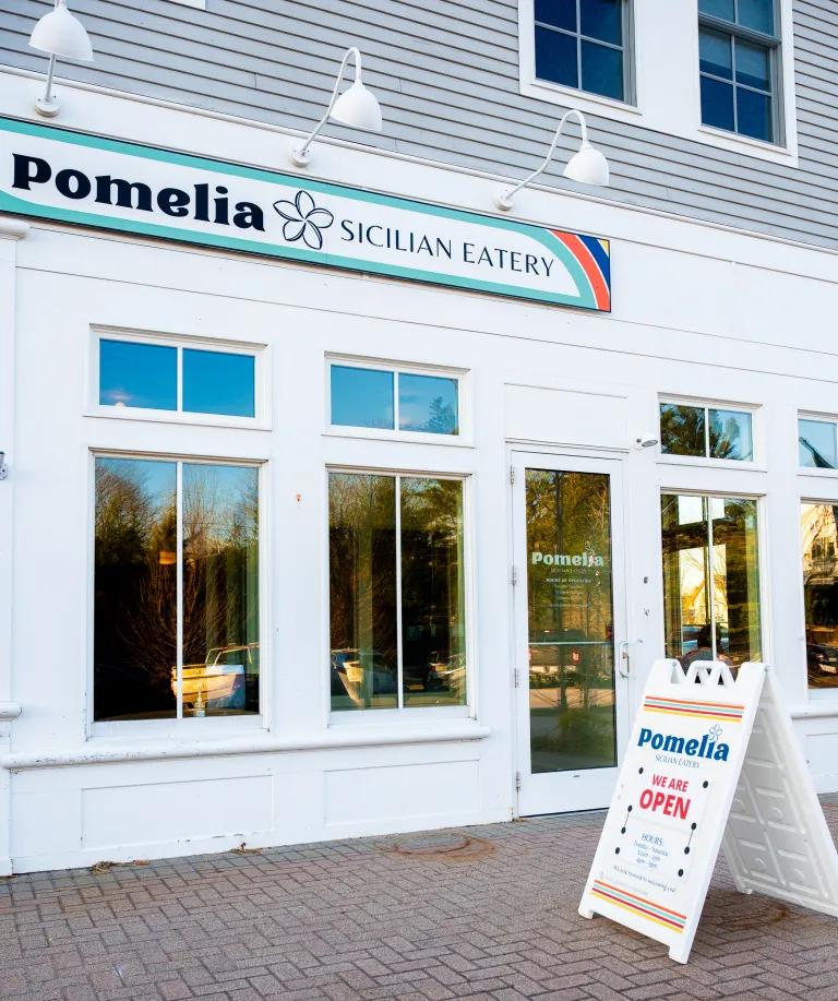 Exterior of Pomelia Sicilian Eatery with white facade, large windows, and a sandwich board sign stating the restaurant is open.