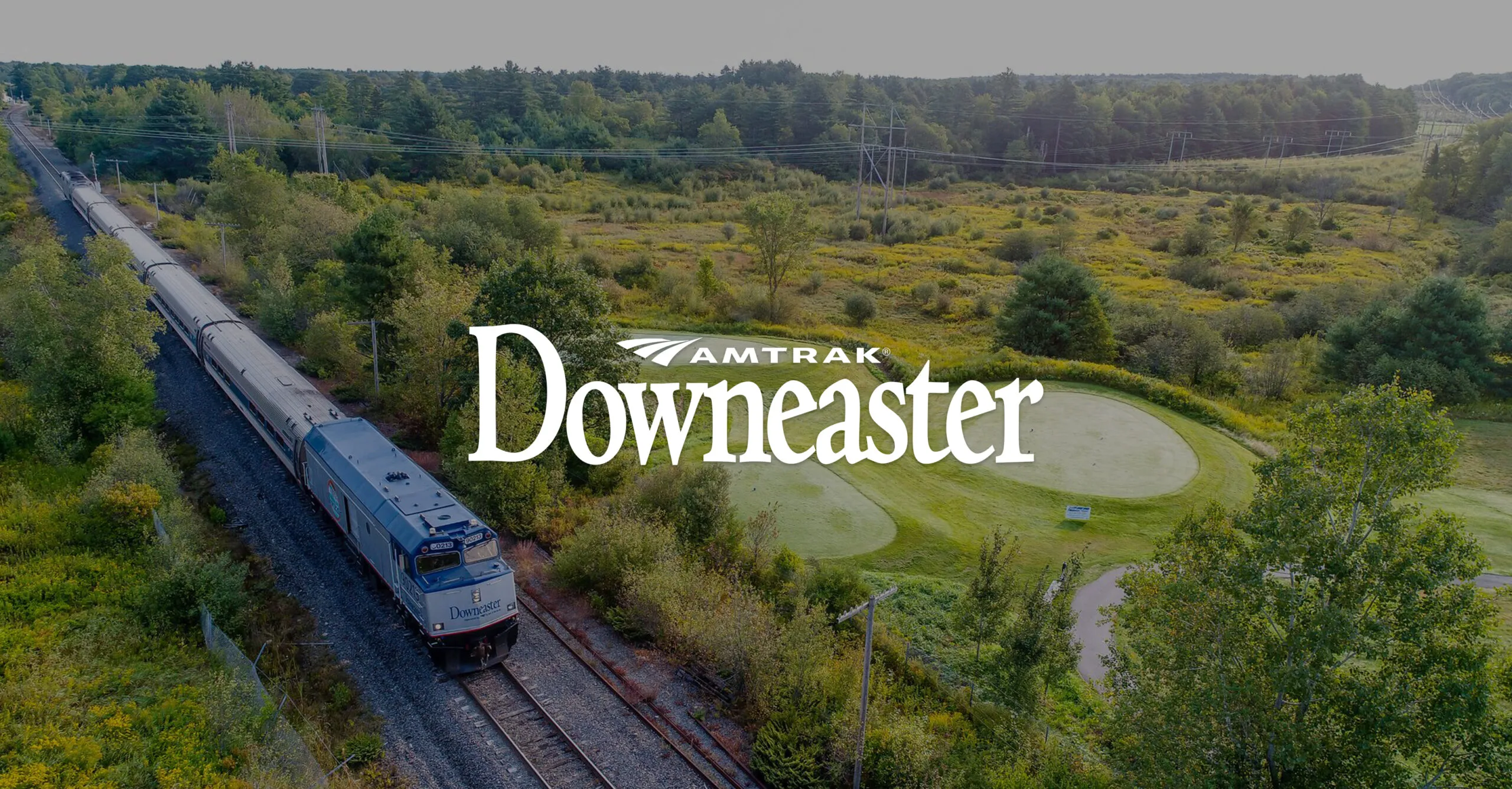 Amtrak Downeaster train traveling on tracks through a green wooded landscape with golf course putting greens nearby.