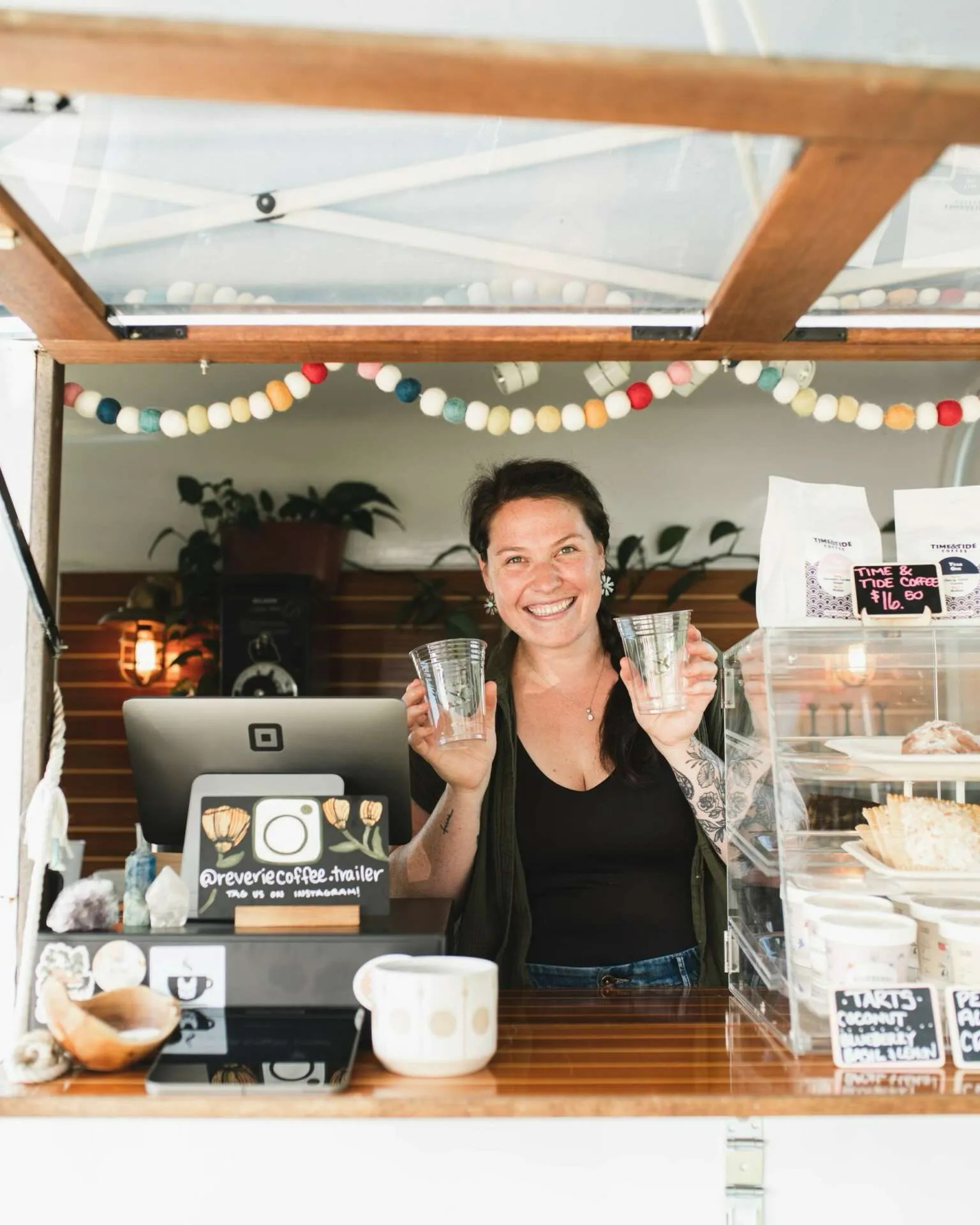 Hannah Josselyn barista behind coffee stand holding two clear plastic cups with decorative garland overhead.