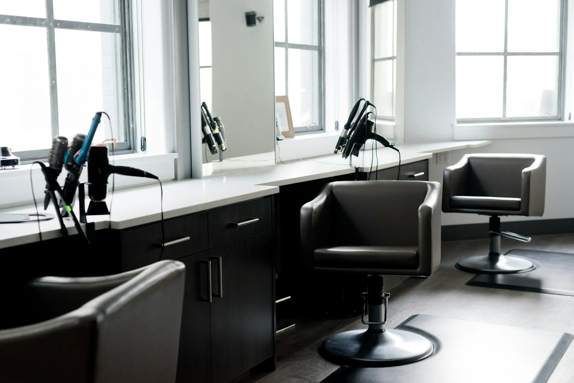 Modern hair salon interior with black styling chairs, countertop, hair dryers, and large windows.