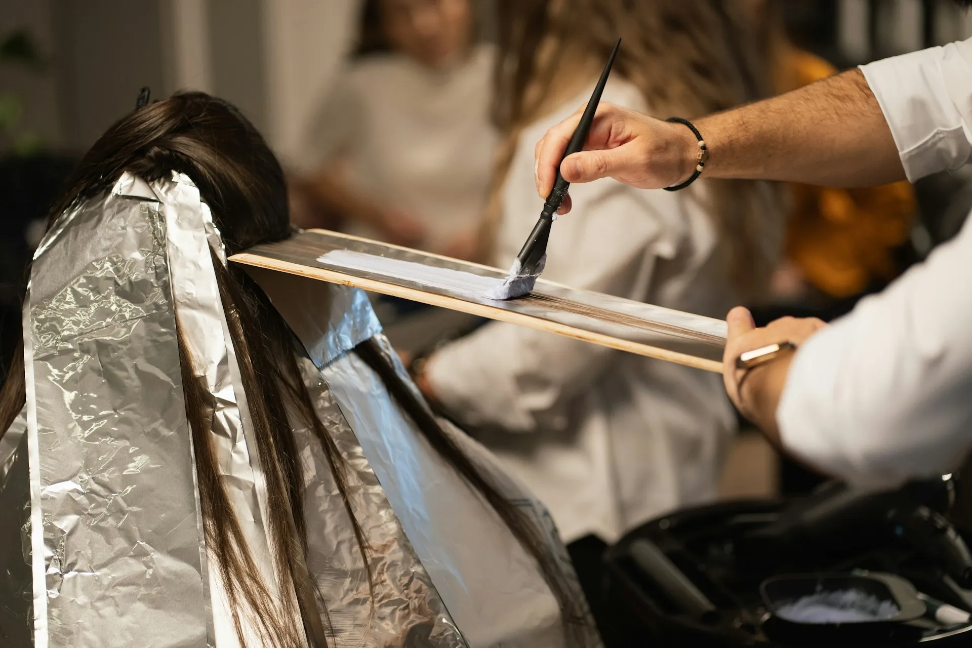 Hairdresser applying hair dye with brush onto a section of hair wrapped in foil at a salon.