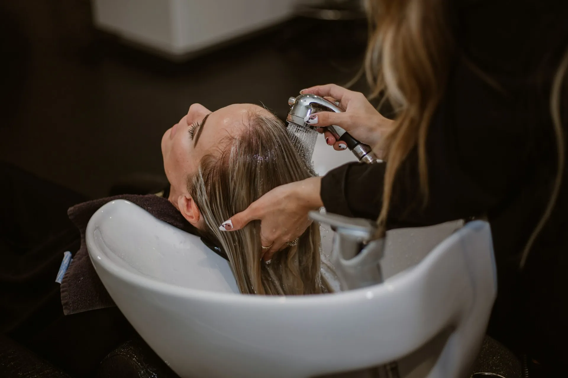 Woman reclining with her hair being washed at a salon sink by a person holding a shower spray.