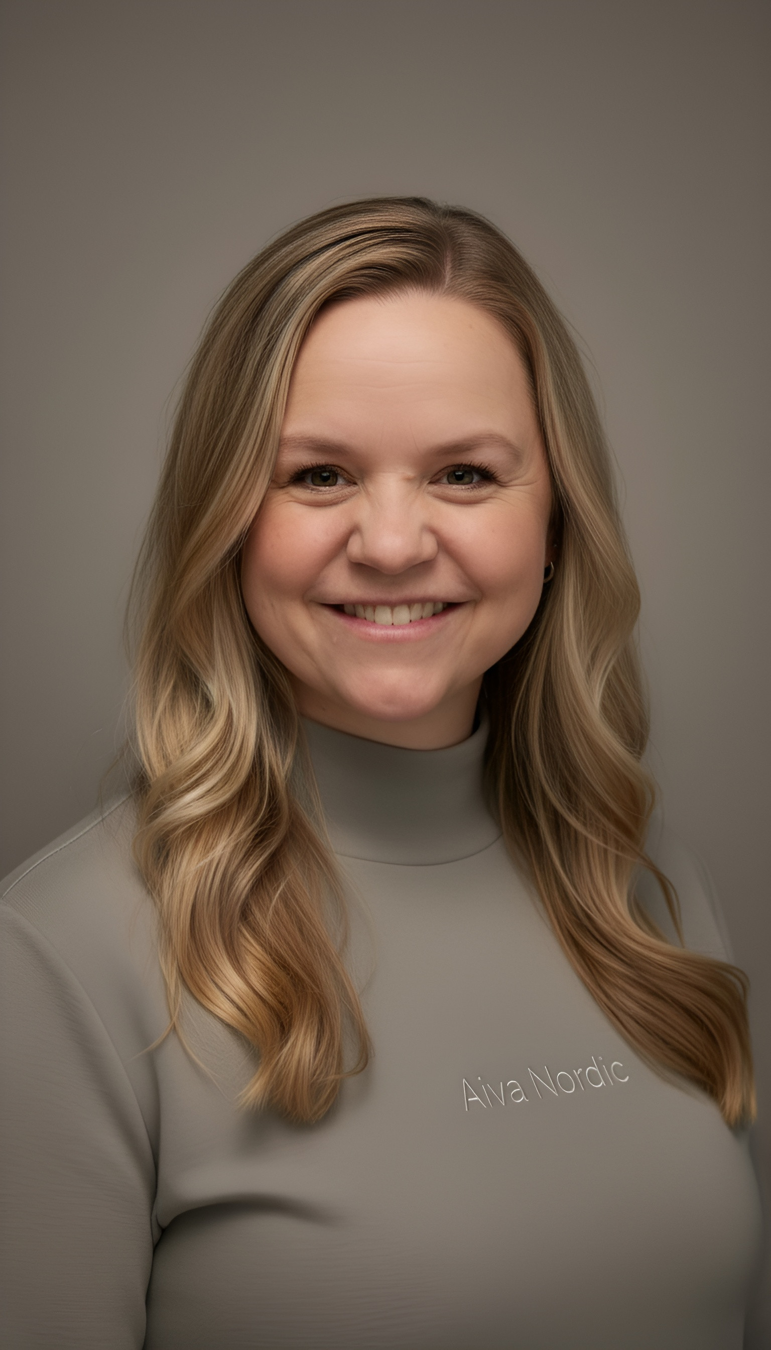 Smiling woman with long blonde hair wearing a dark jacket standing outdoors with a blurred grassy and cloudy sky background.