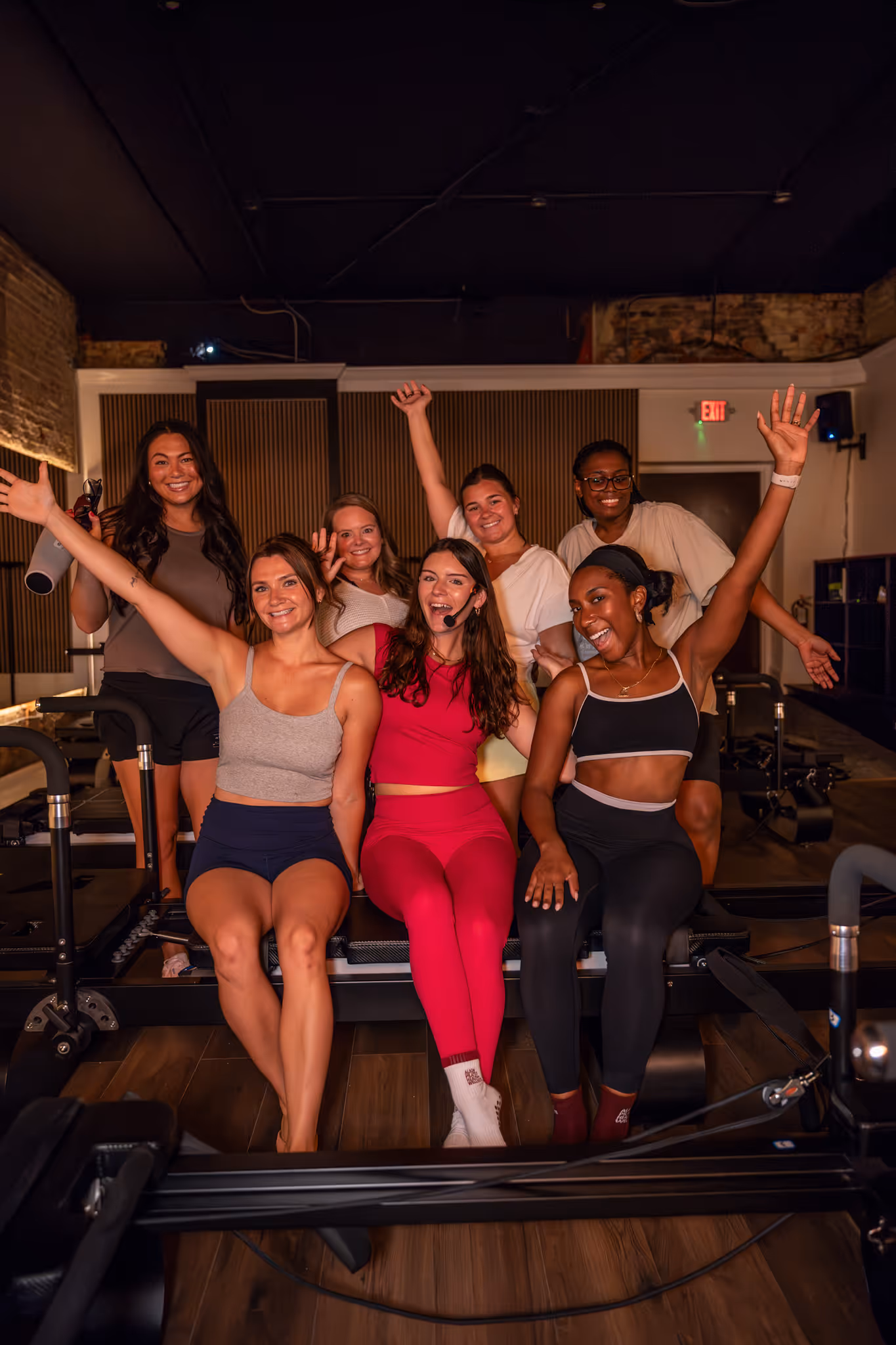 Seven smiling women in workout clothes posing joyfully on fitness reformer machines in a studio.