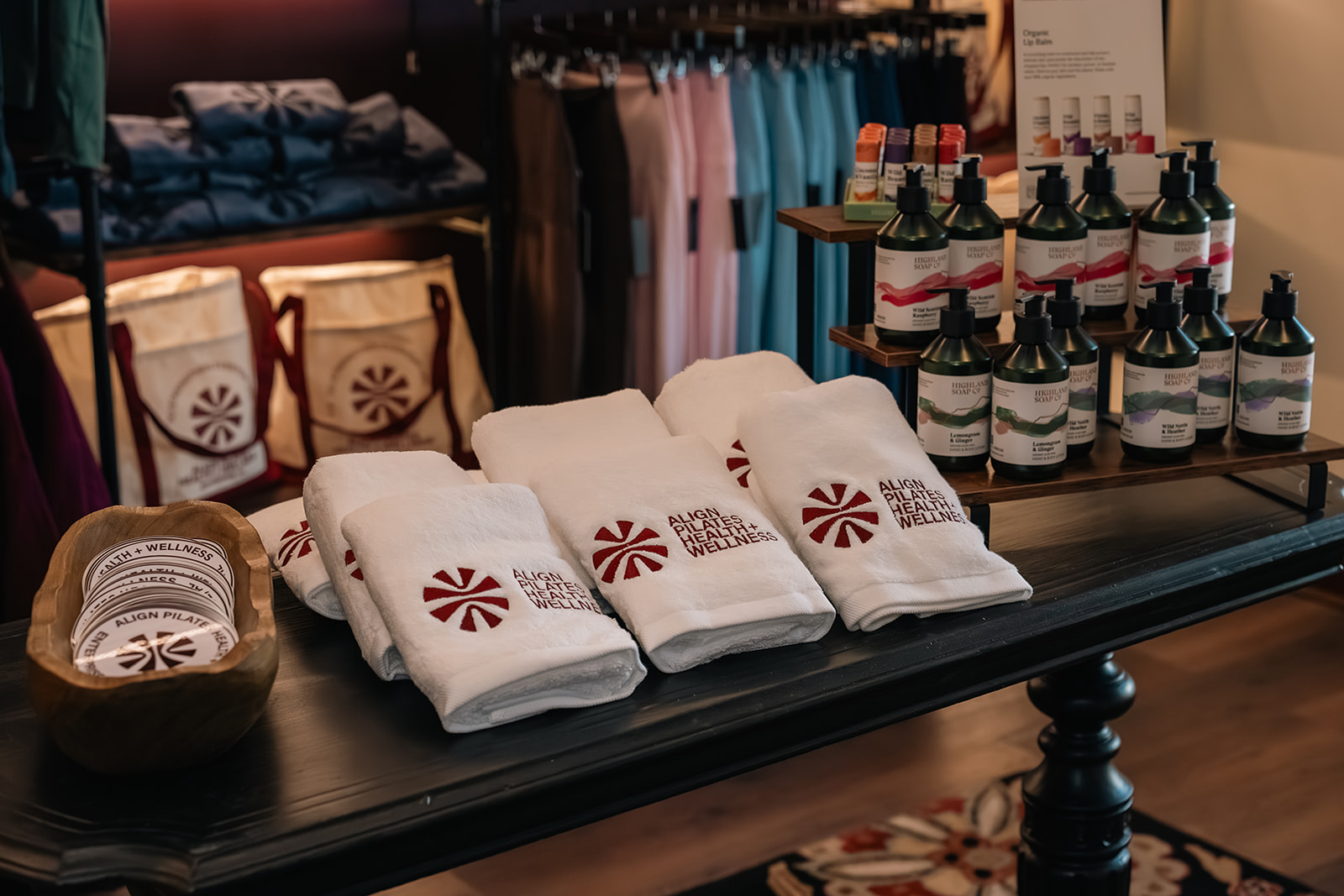 White tote bag with 'The Pilates Class' text, black cap, white bottle, and brown coffee cup on a white couch.