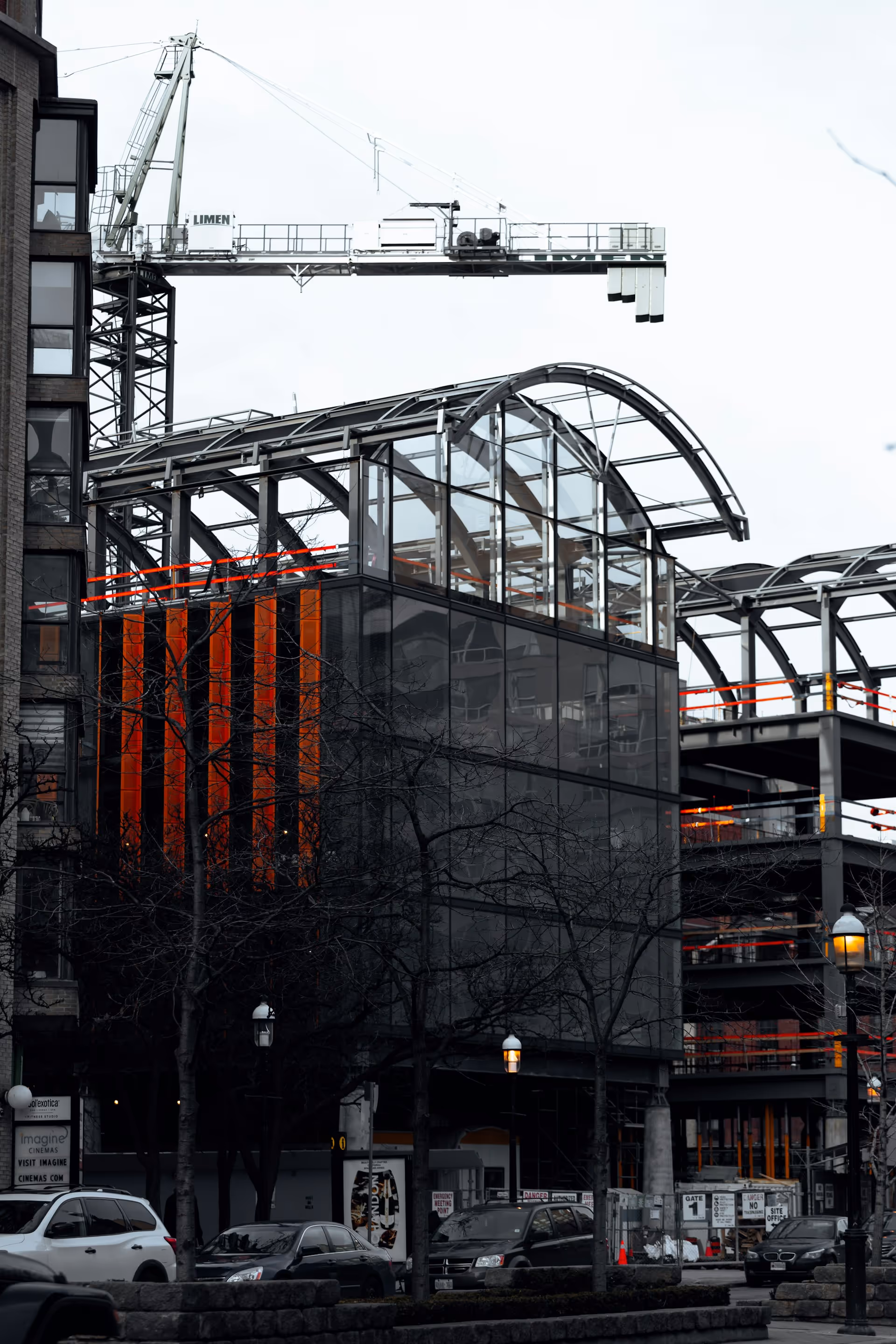 Modern building under construction with glass and metal framework, crane overhead, and parked cars in front.